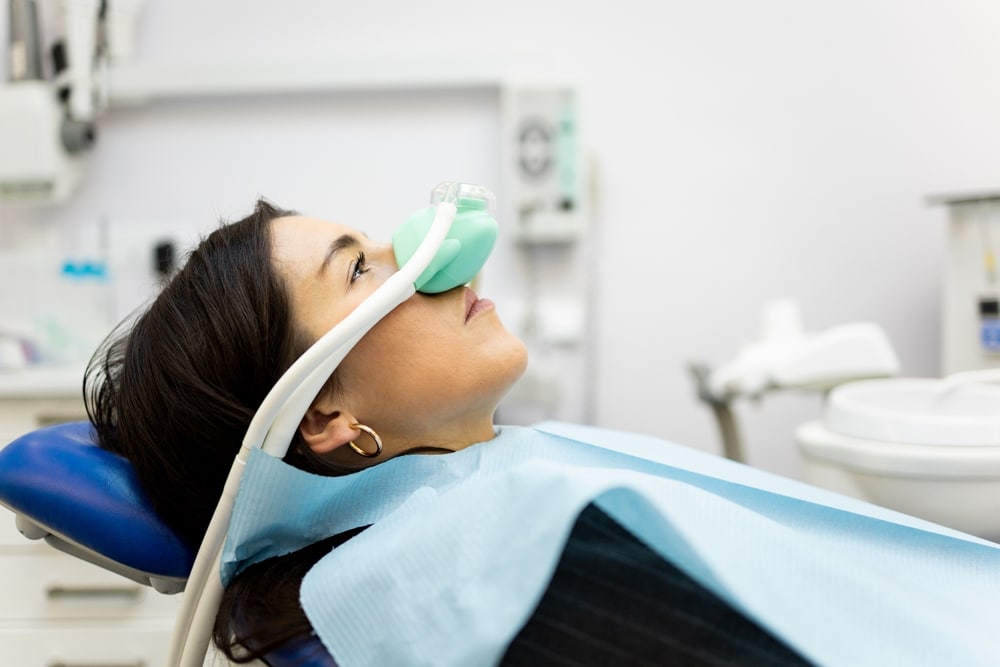 An adult woman sitting in a dentist's office wearing a nasal mask to inhale nitrous oxide.